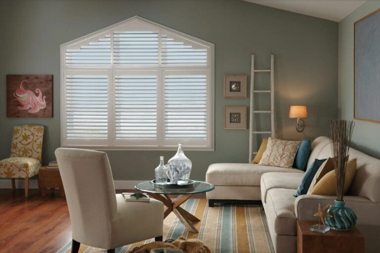 High-End Plantation Shutters in a coastal-themed living room featuring white shutters on an angled window, sage green walls, and modern furniture