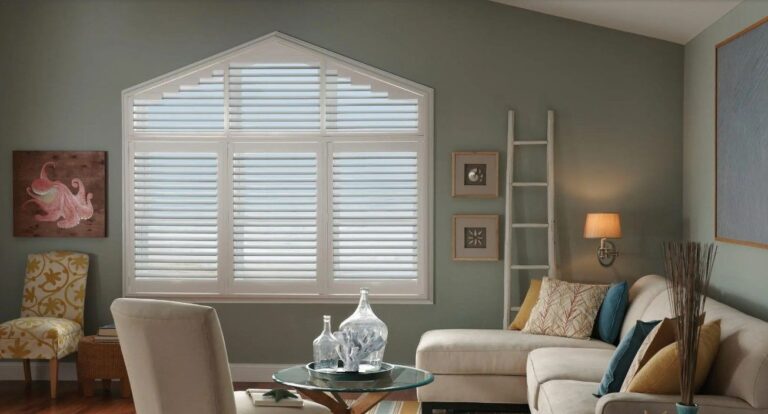 Elegant window treatments featuring white plantation shutters on an angled window, complementing a sage green living room with coastal decor