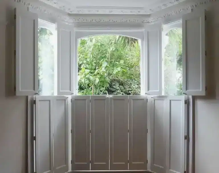 Custom shape shutters enhancing a window with greenery outside, crafted by North London Shutters and Blinds.