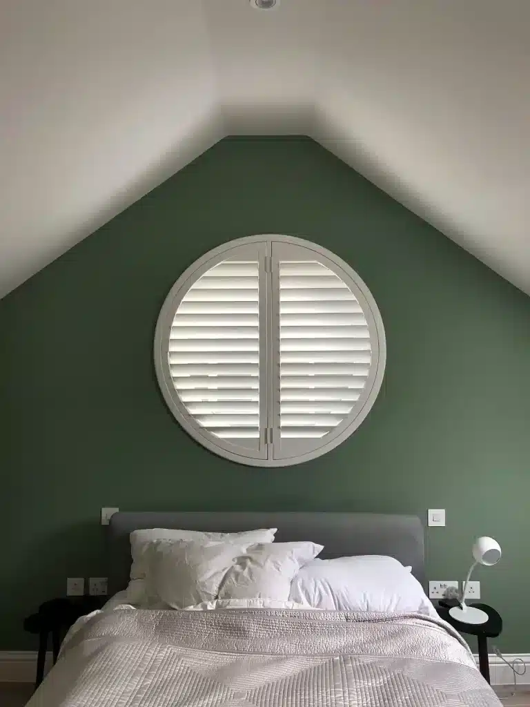 Elegant wooden interior shutters on a circular window in a sage green bedroom, showcasing North London Shutters and Blinds craftsmanship