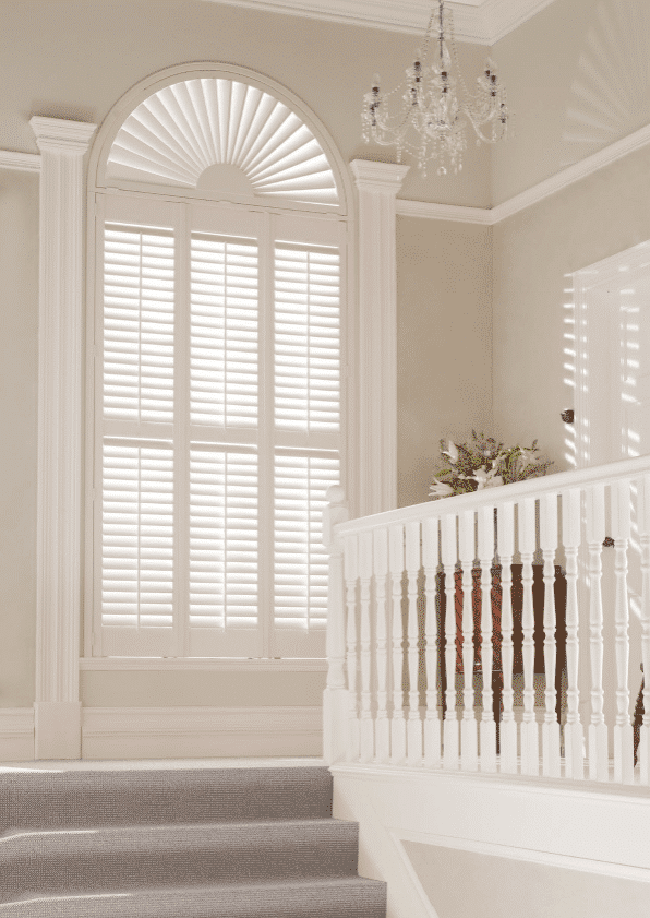 Custom interior wooden window shutters with sunburst arch design installed in a bright stairway landing, showcasing premium plantation-style window treatment