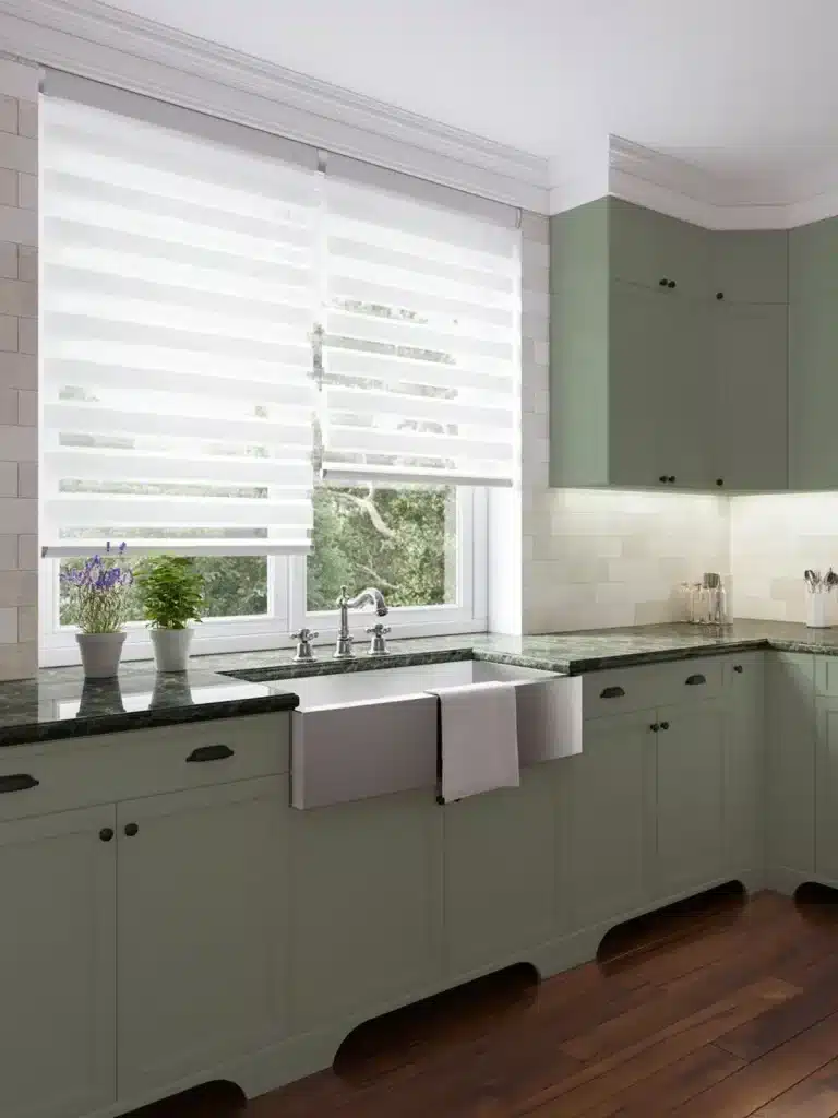 Kitchen interior featuring john lewis shutters style white horizontal blinds above farmhouse sink with sage green cabinets