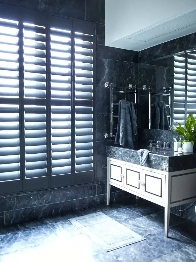 Custom white plantation window shutters in London luxury bathroom with dark marble tiles and modern vanity