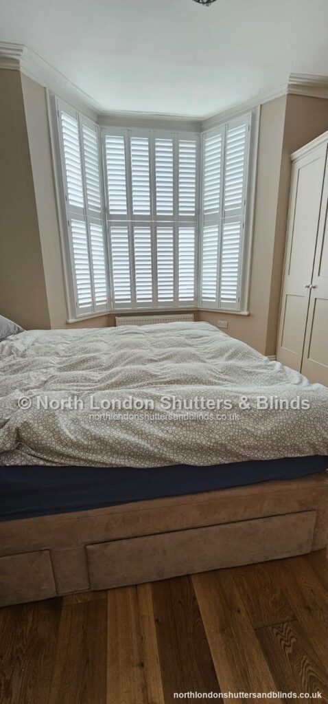 White tier-on-tier plantation shutters on a bay window in a bedroom, with the shutters open by shutter fitters in Barnet, North London