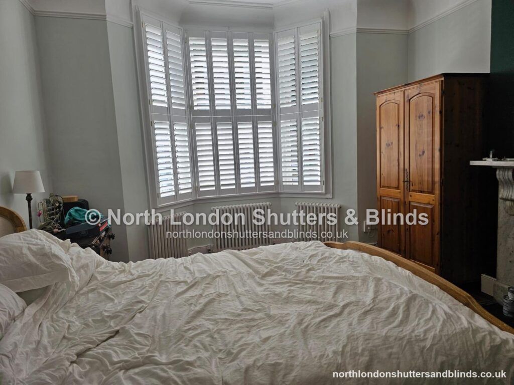 White tier-on-tier plantation shutters on a bay window in a bedroom, with the louvres closed by shutter fitters in Barnet, North London