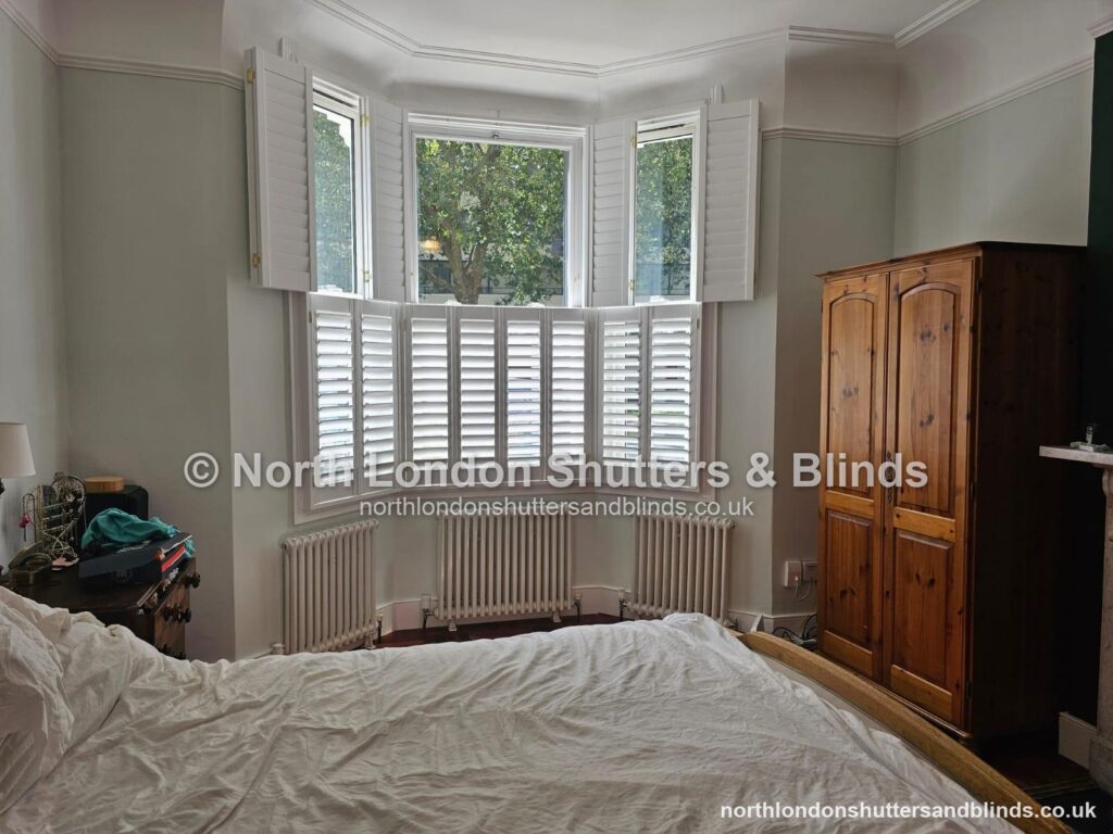 White tier-on-tier plantation shutters on a bay window in a bedroom, with the top panels folded open by shutter fitters in Barnet, North London