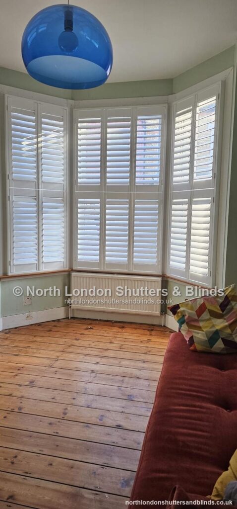 White tier-on-tier plantation shutters on a bay window in a living room, with the louvres closed by shutter fitters in Barnet, North London
