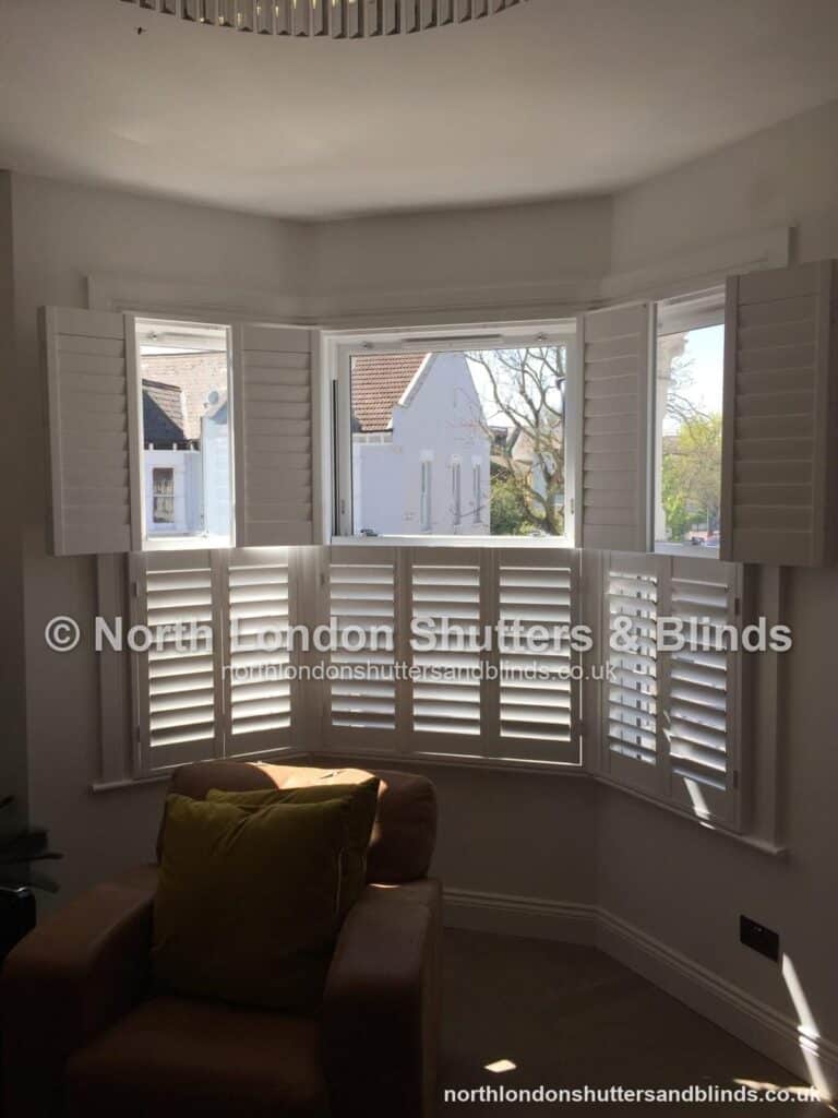 White tier-on-tier plantation shutters on a bay window in a living room, with the top panels folded open by shutter fitters in Barnet, North London